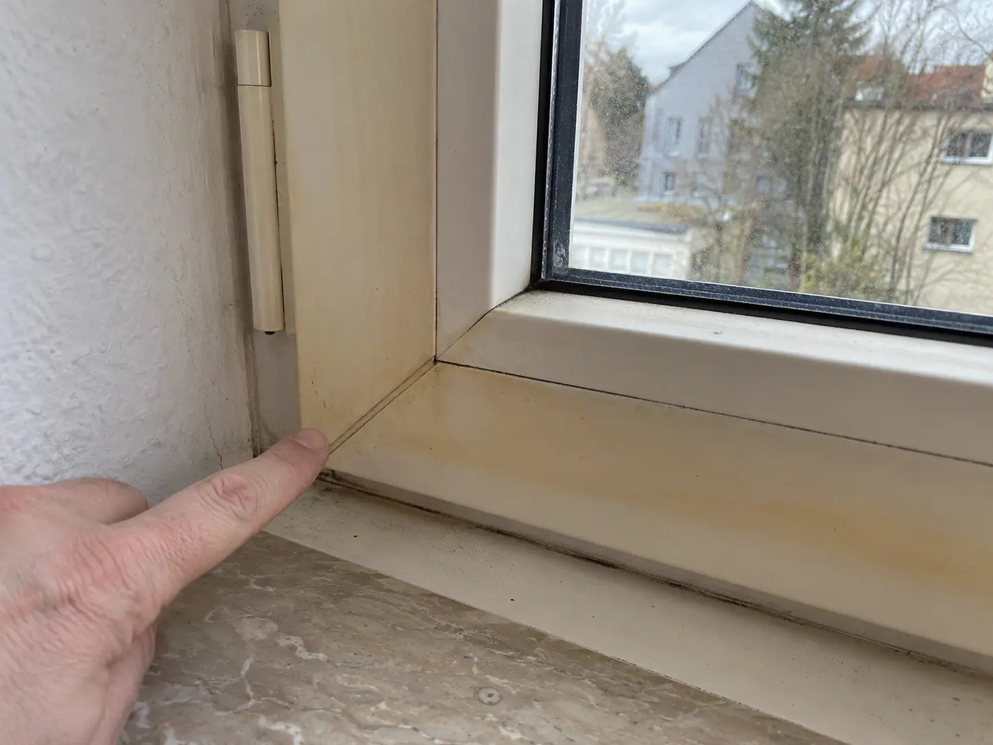 Finger zeigt auf gelblichen Schmutzfilm in der Ecke eines Kunststofffensterrahmens